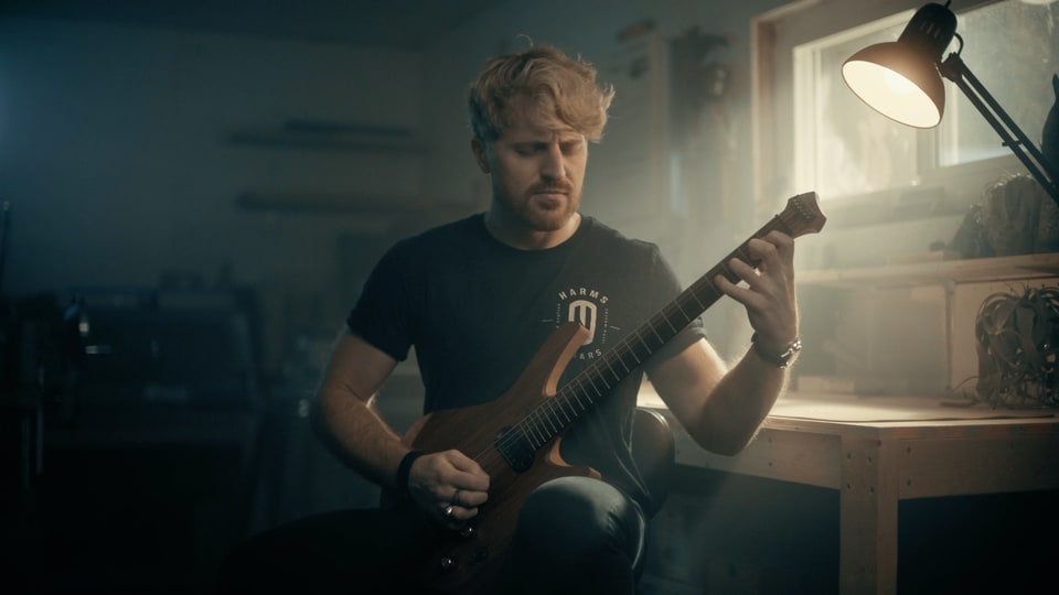 Saul performing Angular in a warmly lit workshop, seated with his guitar beside a window.