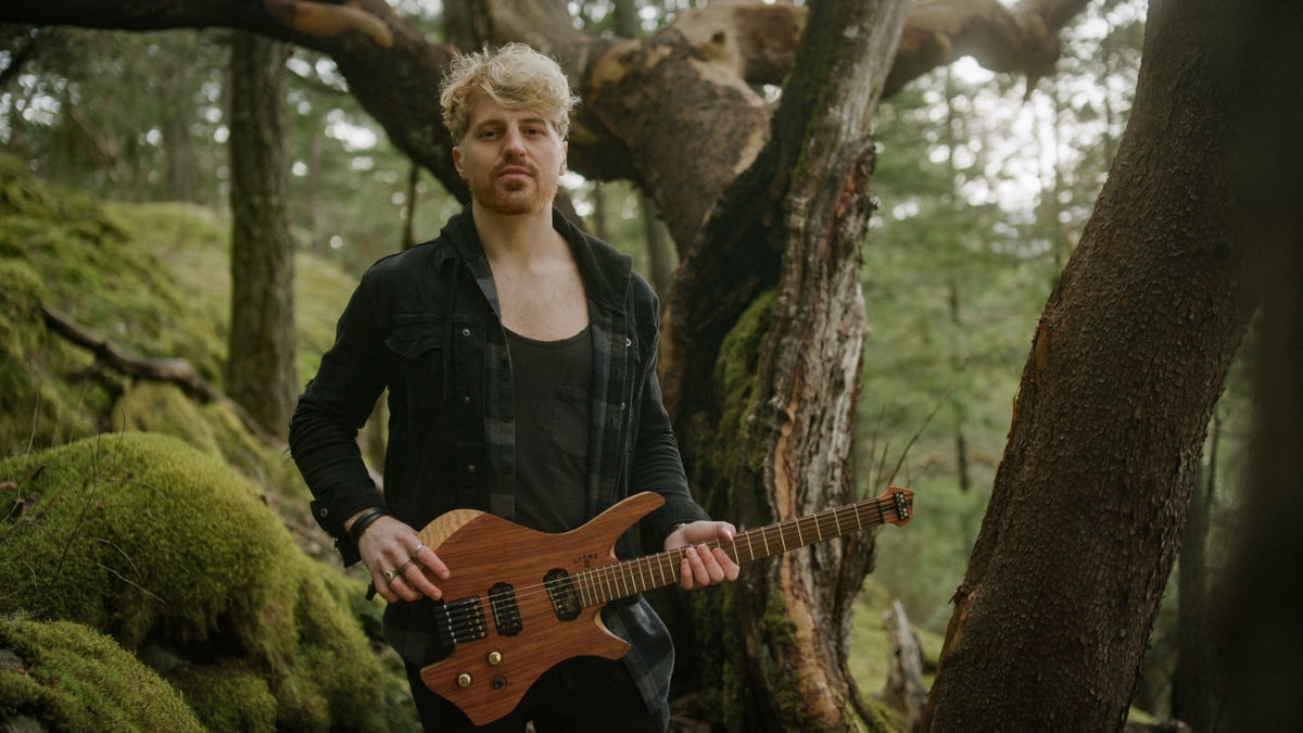 Saul holding a sculptural wood-bodied guitar among moss-covered trees.