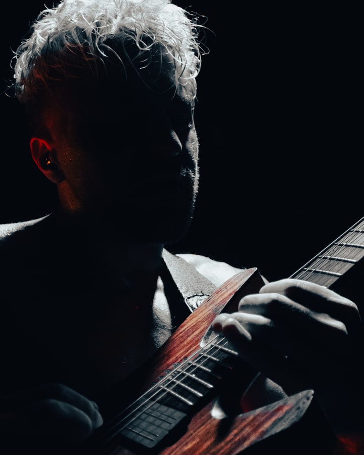 A quiet, rim-lit portrait of Saul, with his hand and wood guitar emerging from shadow.