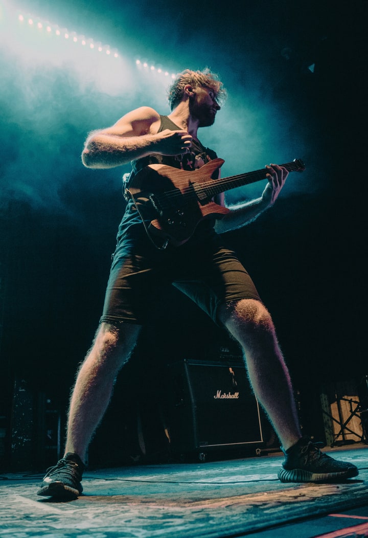 Saul Sitar playing his wood-bodied guitar in a wide, grounded stance beneath cyan stage light.